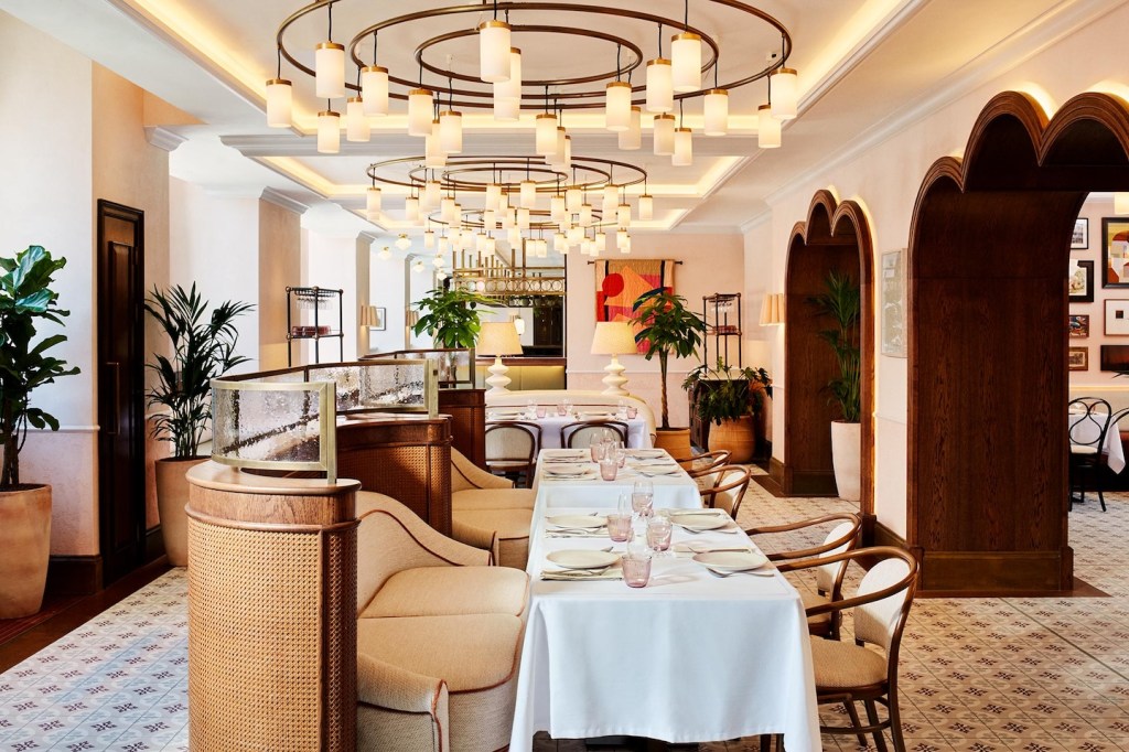 A long view of the restaurant's main dining hall, featuring multiple circular brass chandeliers with cylindrical white bulbs. The room is filled with booths and tables set for service, flanked by tall potted plants and decorative wooden arches.